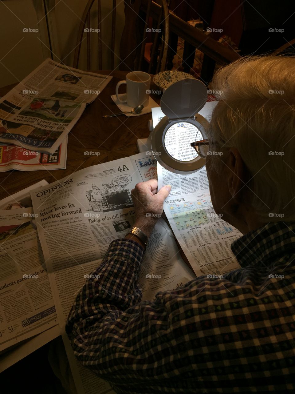 Elderly senior citizen male using lighted electronic magnifying glass gadget to read newspaper print while seated at kitchen table.