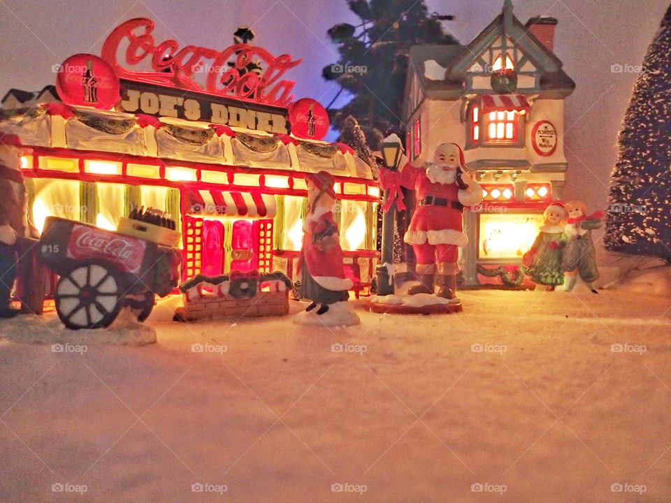 A Christmas village display with a Coca Cola sign on top of Joe’s Diner and Santa Claus with people out front. 