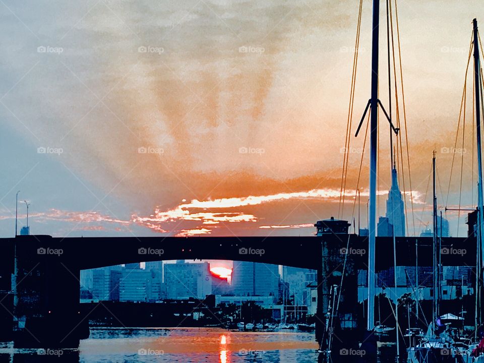 Sunset over the Pulaski Bridge at Long Island City’s Newtown Creek in Queens, NY with sailboat. The sun’s dying rays cast ripples resembling the spikes of a crown into the air as well as intense orange hues. Photo from 2018. Hypnotic Productions