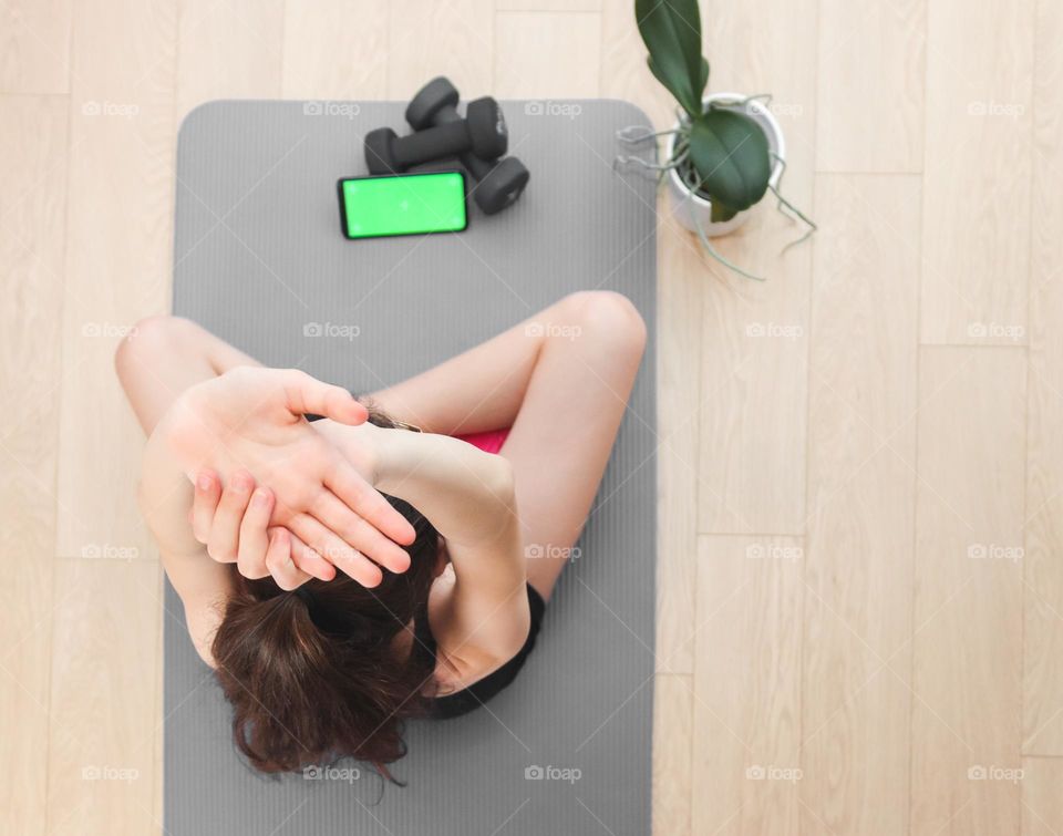 One young caucasian girl sits on a mat on the floor in sportswear cross-legged and stretched her arms up, looks into a smartphone with a green screen on black dumbbells and repeats a fitness exercise, top view close-up.