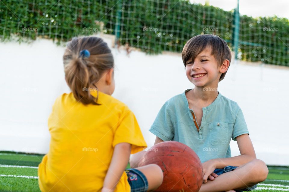 Positive boy sitting near girl and talking with her while spending time together on green lawn during break from football game