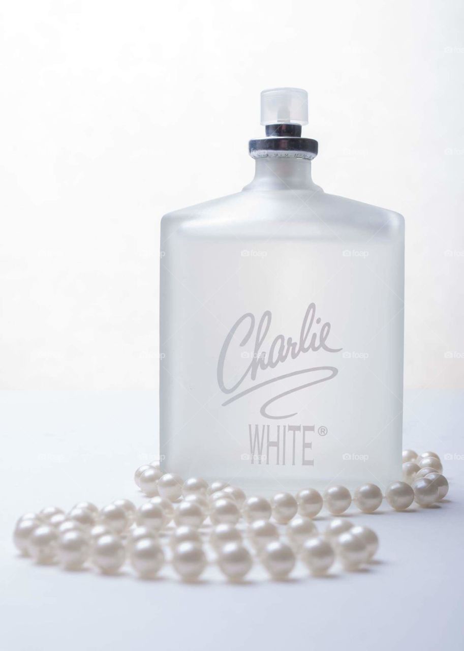all white on white image of white frosted perfume bottle wrapped by a string of white pearls on a white table with a white background