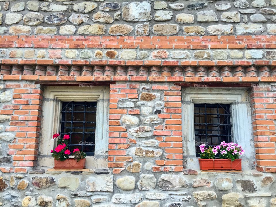 Two old looking windows with bars decorated with flower pots on a wall with bricks