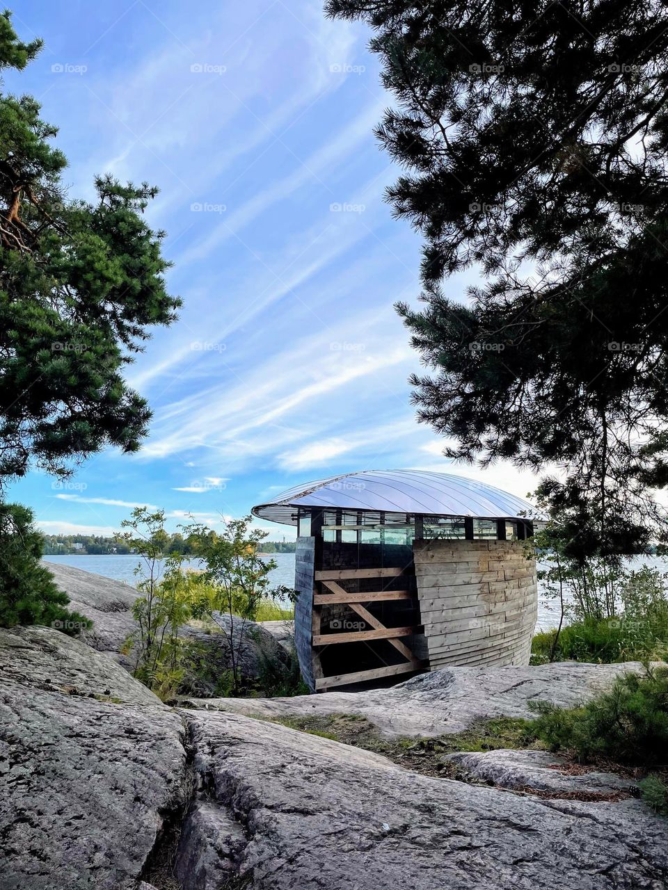 View to deep blue sky with white cloud lines from the rocky island with green trees and futuristic shape wooden with plexi plastic roof boathouse