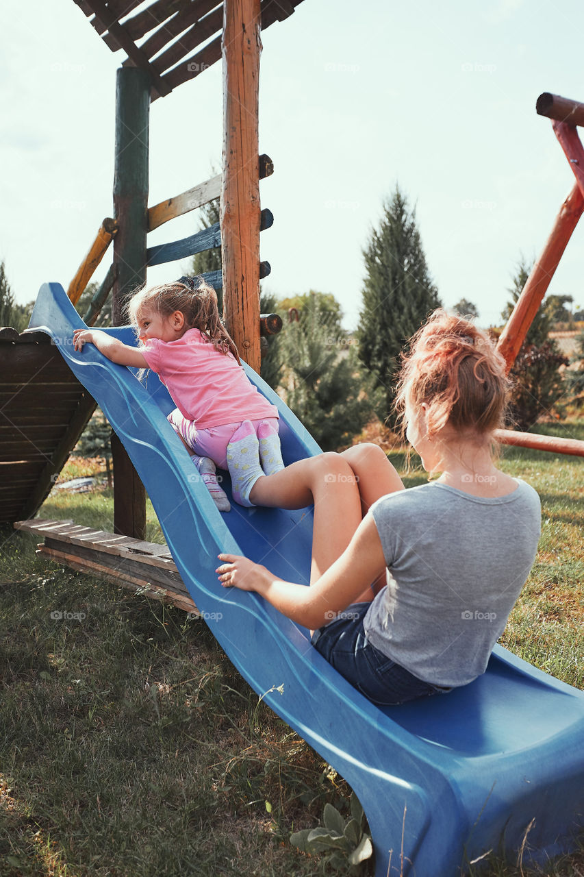 Teenage girl playing with her younger sister in a home playground in a backyard. Happy smiling sisters having fun on a slide together on summer day. Real people, authentic situations