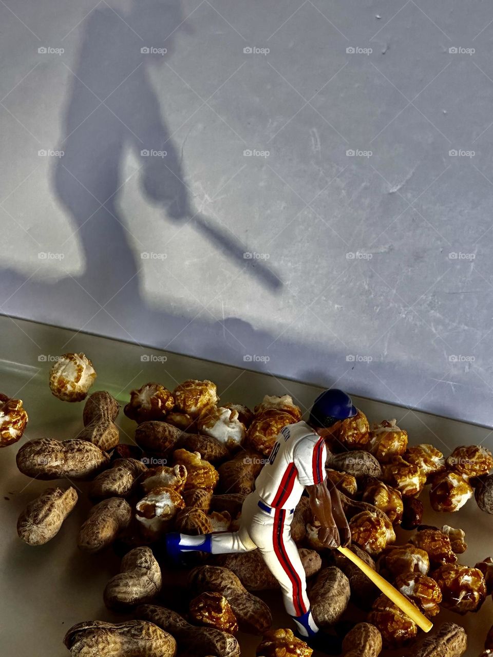 Plastic sports action figure baseball slugger Darryl Strawberry in Mets uniform standing in actual peanuts and Cracker Jack in front of his shadow cast on a foam core board background 