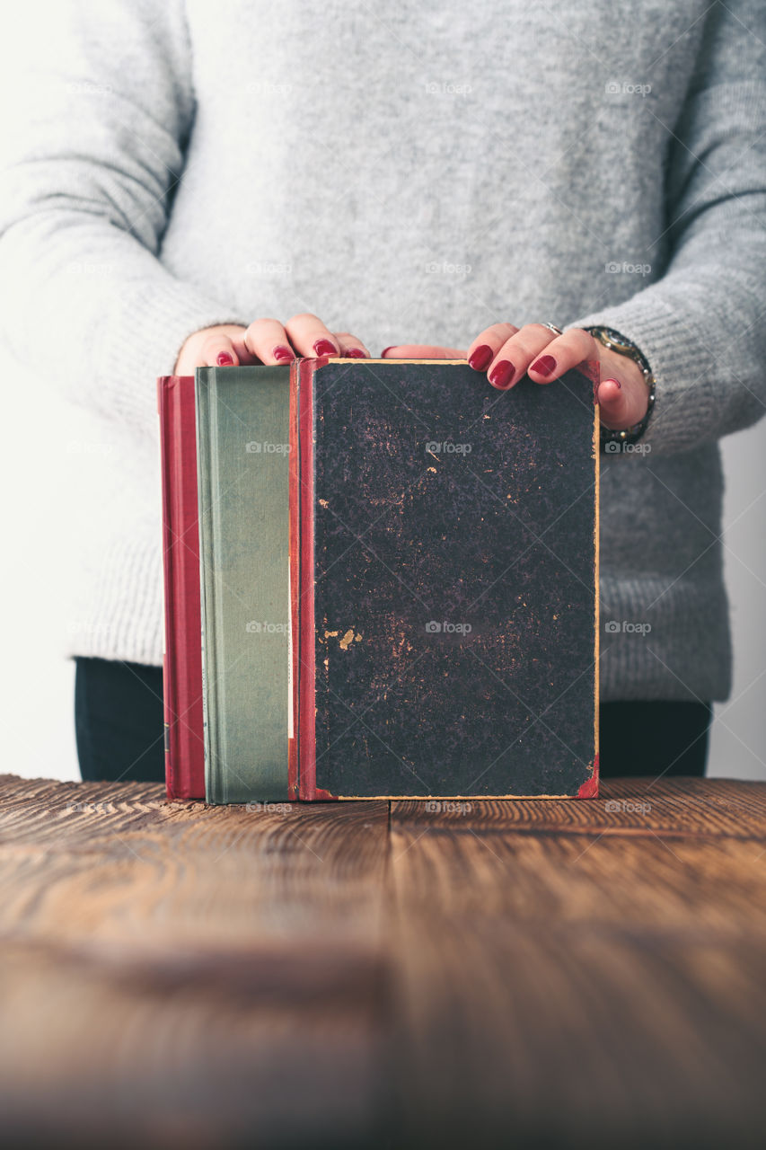 Young woman holding a few old books on wooden table in antique bookstore. Woman wearing grey sweater and jeans. Vertical photo