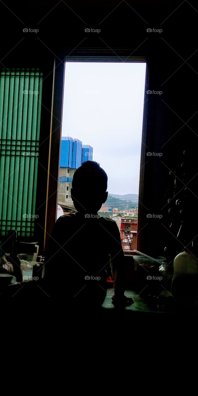 the silhouettes of a boy sitting on the table turning his back from the window.