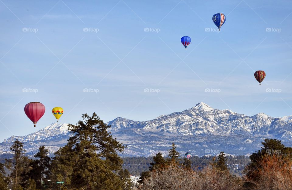 A hot air balloon festival in the winter creates a colorful scene. A forest and mountain make the shot.