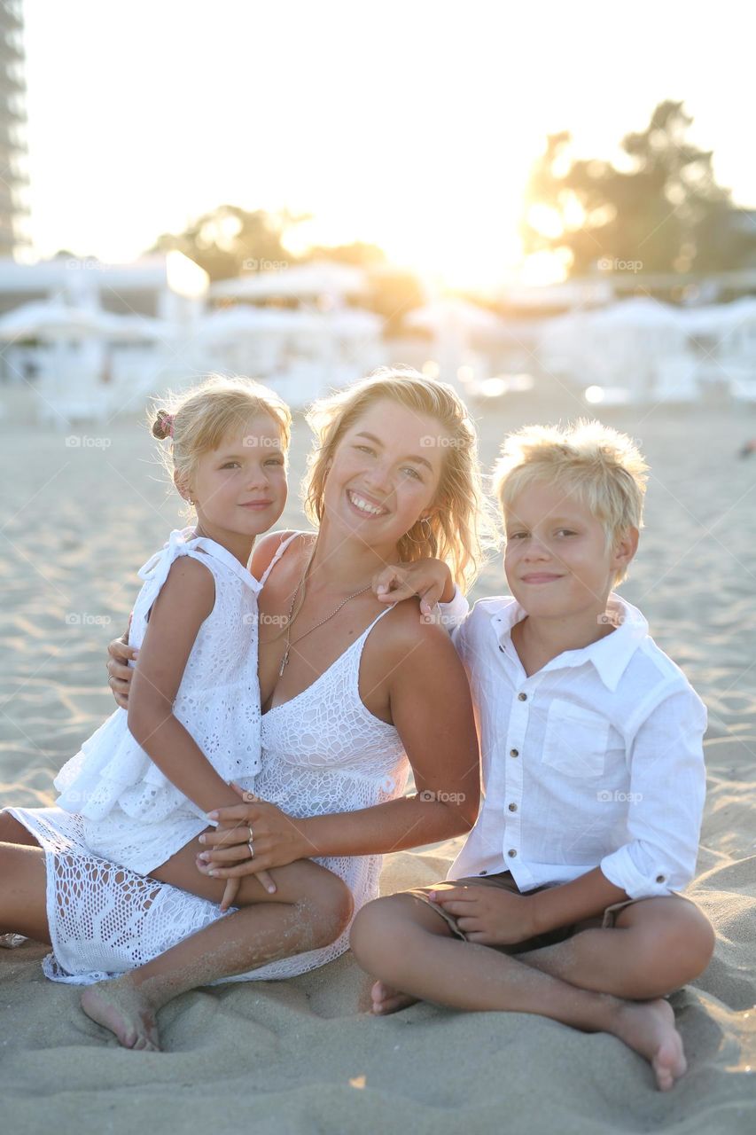 beautiful mother and her children, all dressed in white clothes, the family is sitting on the beach