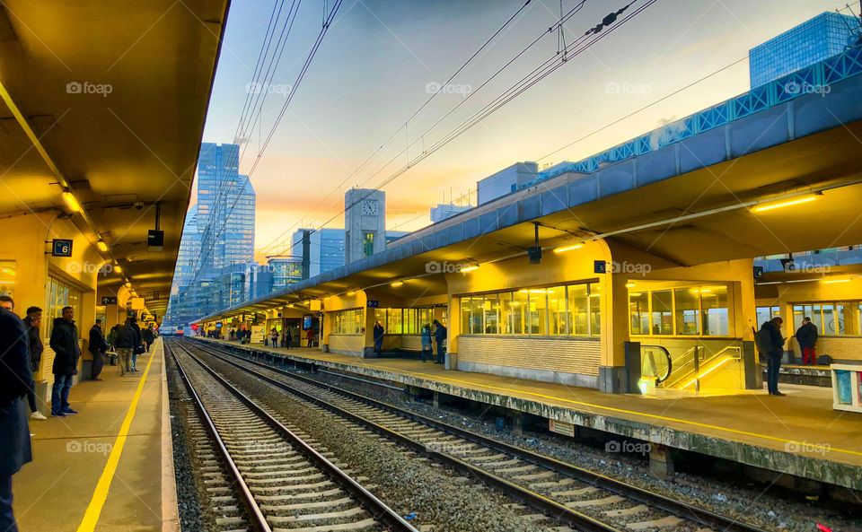 Commuters waiting for the train on the platform of a railway station in the city under a sunset sky