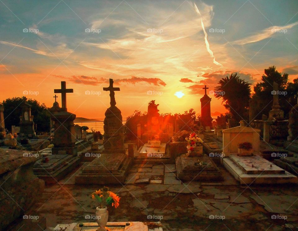 Vivid sunset with dark silhouettes of mausoleums and crosses and orange shaft of sunlight reflecting on orange flowers and sea in the background