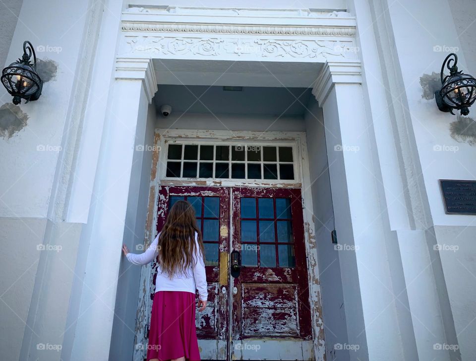 Architectural and historical building main entrance with signs of aging with paint chipped with young girl curious to see how it looks like up the stairs between the white door entrance frame.