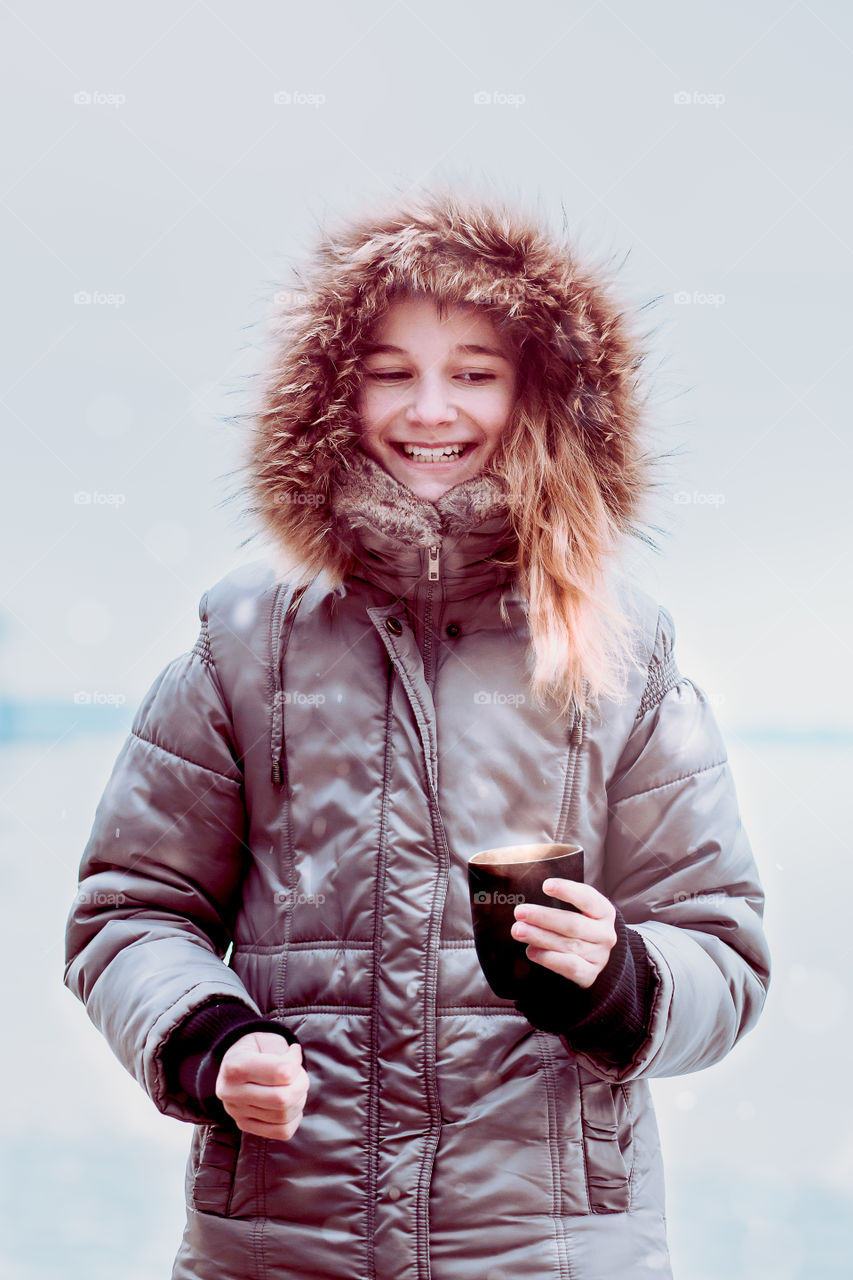 Young pretty smiling girl enjoying drinking hot drink during walk on a wintery day. Wearing coat with hood. Holding a mug of hot tea