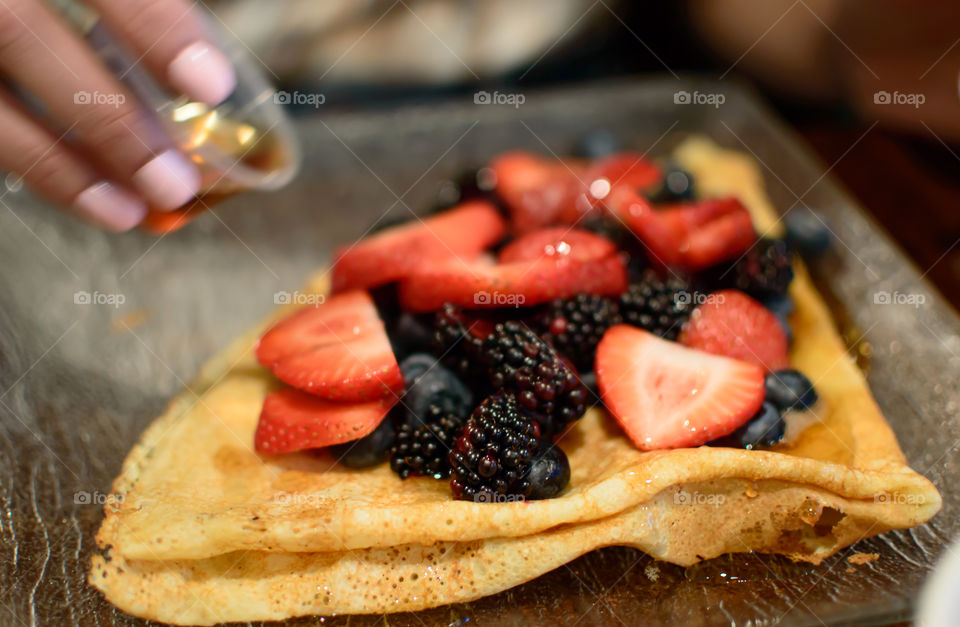 Woman pouring maple syrup on a French crepe with a pile of healthy antioxidant rich raw berries including sliced strawberry, blueberry and blackberry fruit topping 