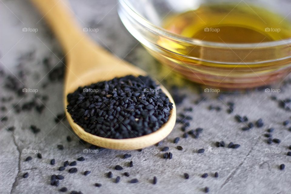 Closeup of black seed in a bamboo spoon next to a small glass bowl of oil on a stone surface