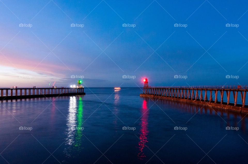 A fishing boat with lights on navigates between two lit piers at dusk