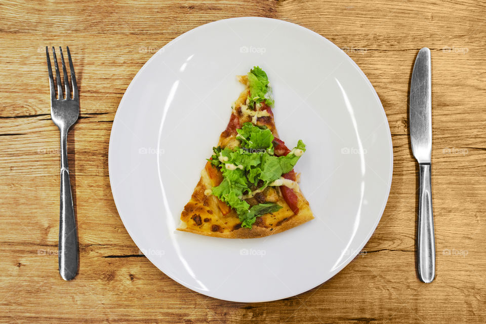 A piece of pizza on white plate with a knife and fork on wooden background. Top view