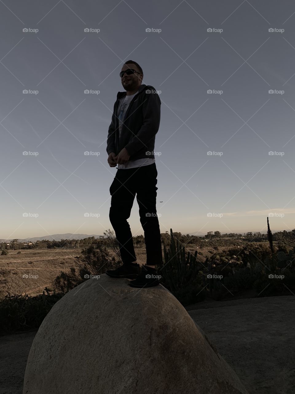 Blonde man standing on desert rock with smile and putting sweater on , sunglasses on , San Diego , California 