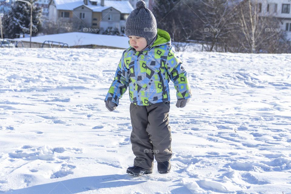 A child in winter in winter jackets, pants, a hat and boots on white snow on the street and in the park in nature plays winter fun and sleds.