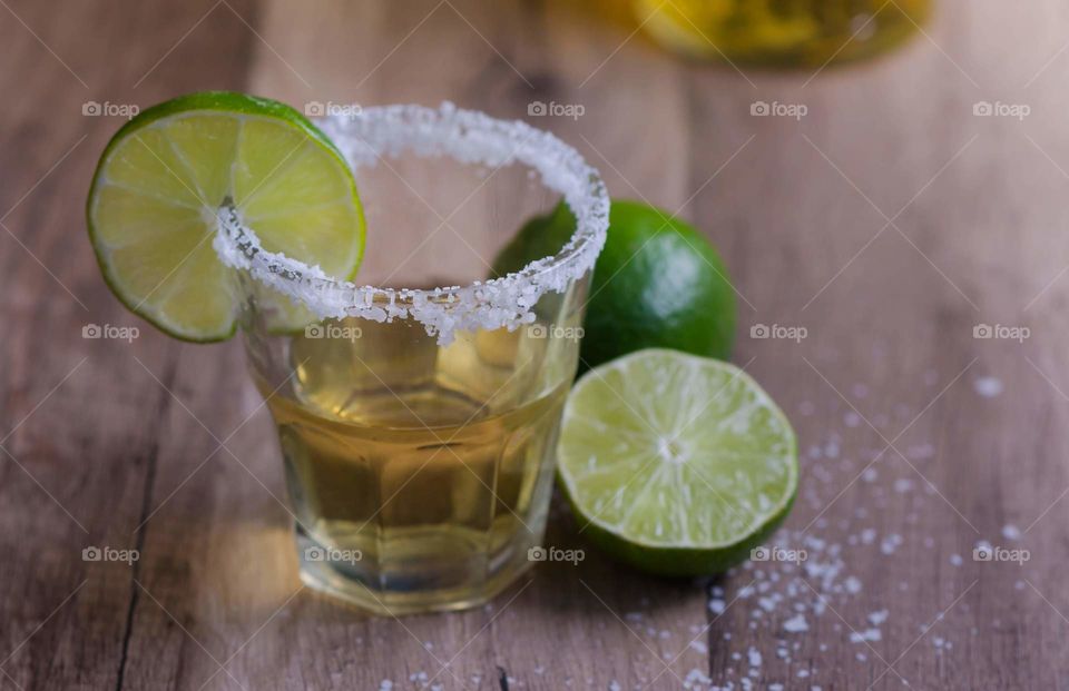 shot of tequila with salt and a lime wedge on the rim sitting on a wood table next to a sliced lime
