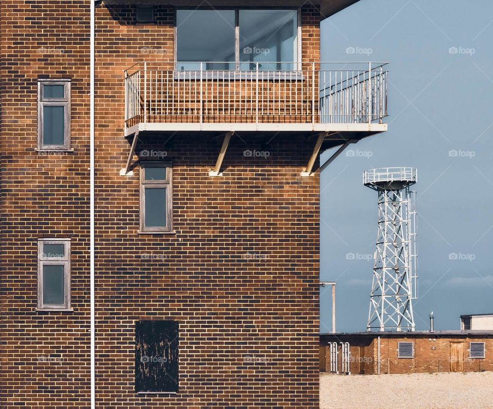 A brick, square building with rectangular windows and a metal tower in the background 