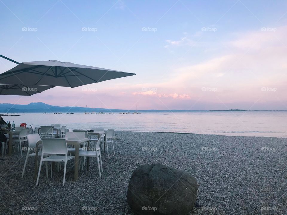 by colorful lake shore at sunset , there are many small stones, rocks , some sets of tables ,chairs. in the purple pink cloudy sky, and the color reflection on the lake surface.  very romantic