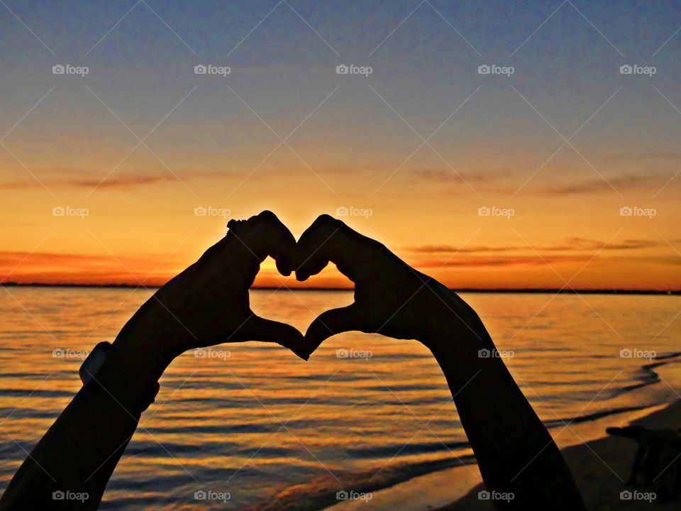 
Living in harmony - Silhouette of woman's hands in heart shape at a spectacular sunset over the bay against a blue-orange sky. Symbol of love and living in harmony