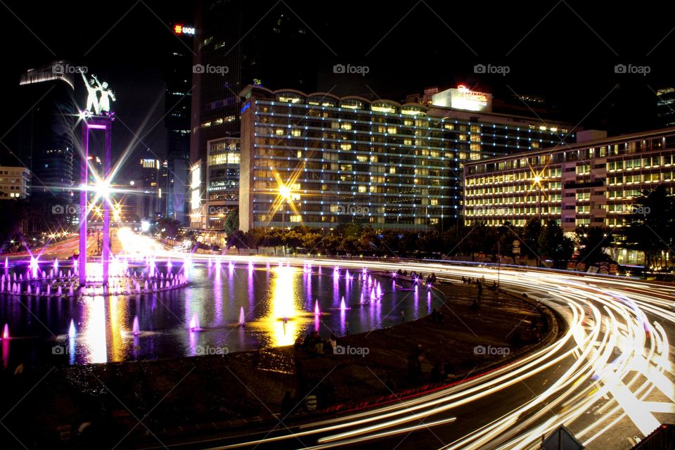 View of the Bunderan HI or Welcome Monument at night in downtown Jakarta with a Hotel and business center building in the background on Jalan Jenderal Sudirman, Jakarta, Indonesia.