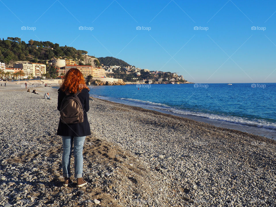 Redhead young woman with backpack walking on the beach in Nice, France.