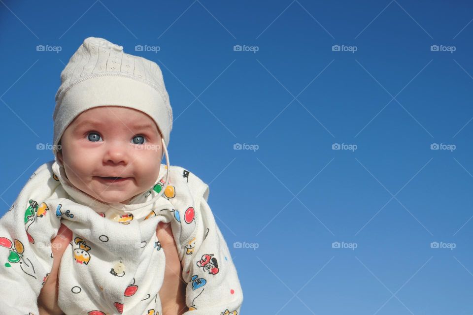 Smiling baby in white clothes and cap on the blue sky background