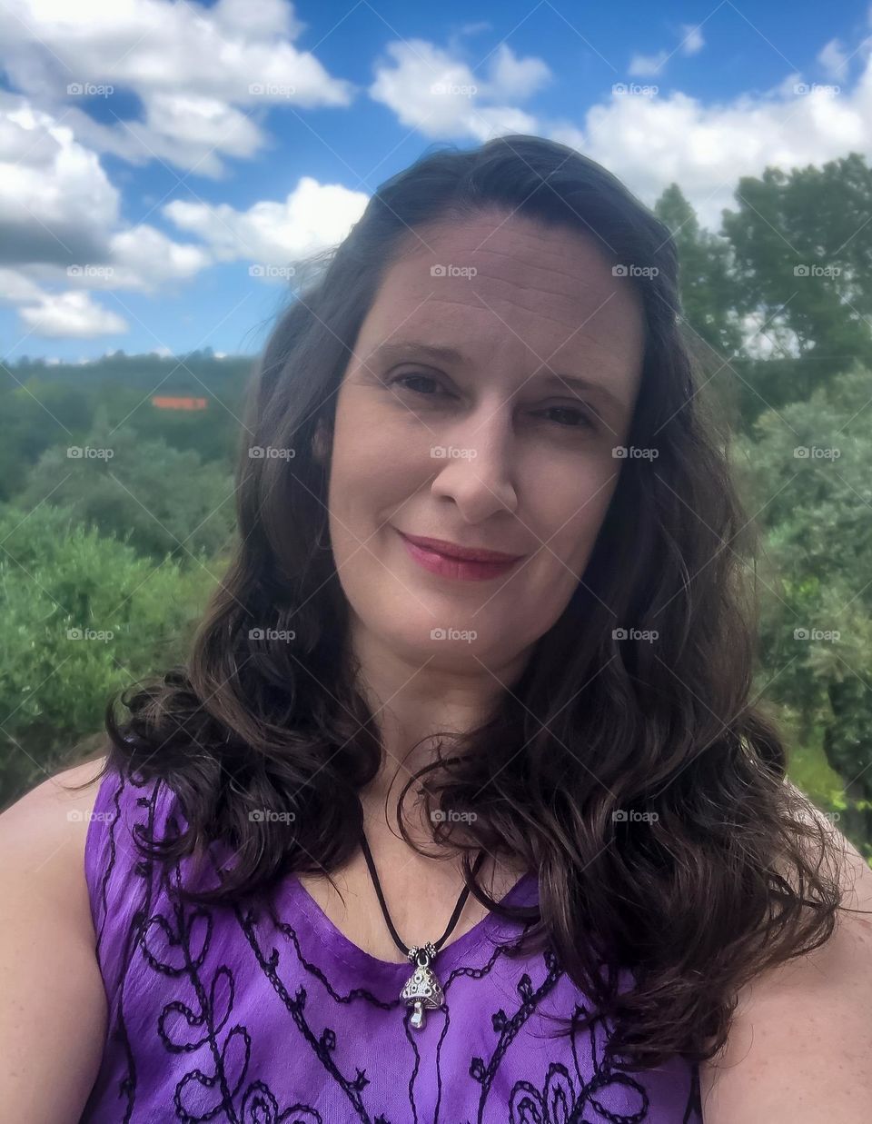 Woman with mid-length dark brown wavy hair against an outdoor background on trees and blue skies