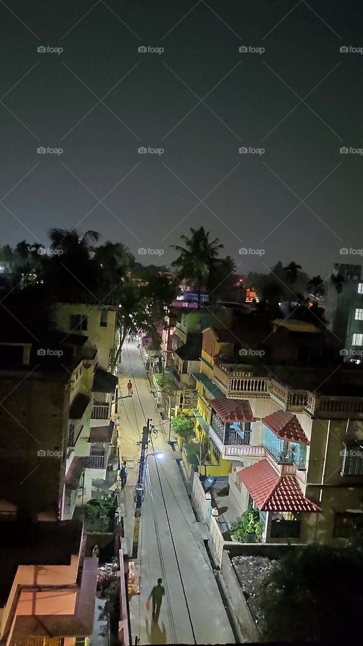 This is a photo of night urban life. The image has been taken from a high-rise building. Night sky high-rise buildings, roads, and silhouettes of trees are shown.