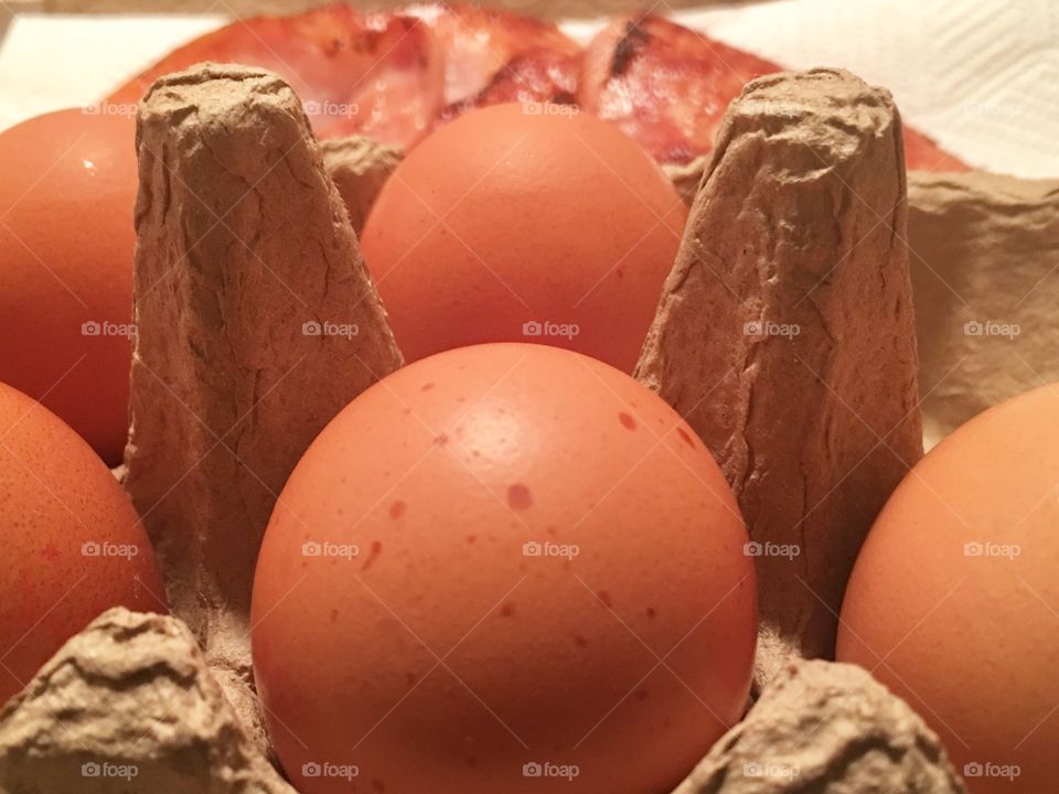 Brown speckled chicken eggs in box in foreground back bacon in background closeup 