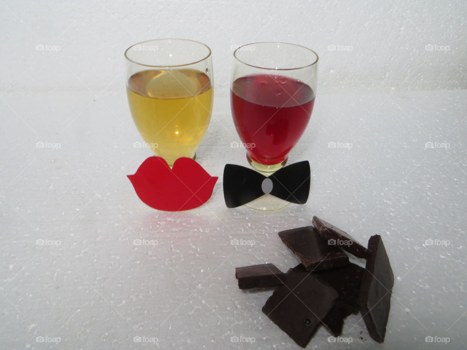 On a white background, two glasses from the left white wine with red lips to the right red wine with a butterfly and black chocolate broken into pieces