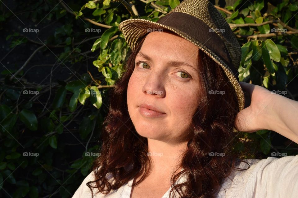 Women with natural and minimal makeup standing outdoors outside with wet hair. Women with red hair and green eyes standing next to tree taking a selfie. Sunlight shine on women hair and skin and making shadows. Women wearing a hat.
