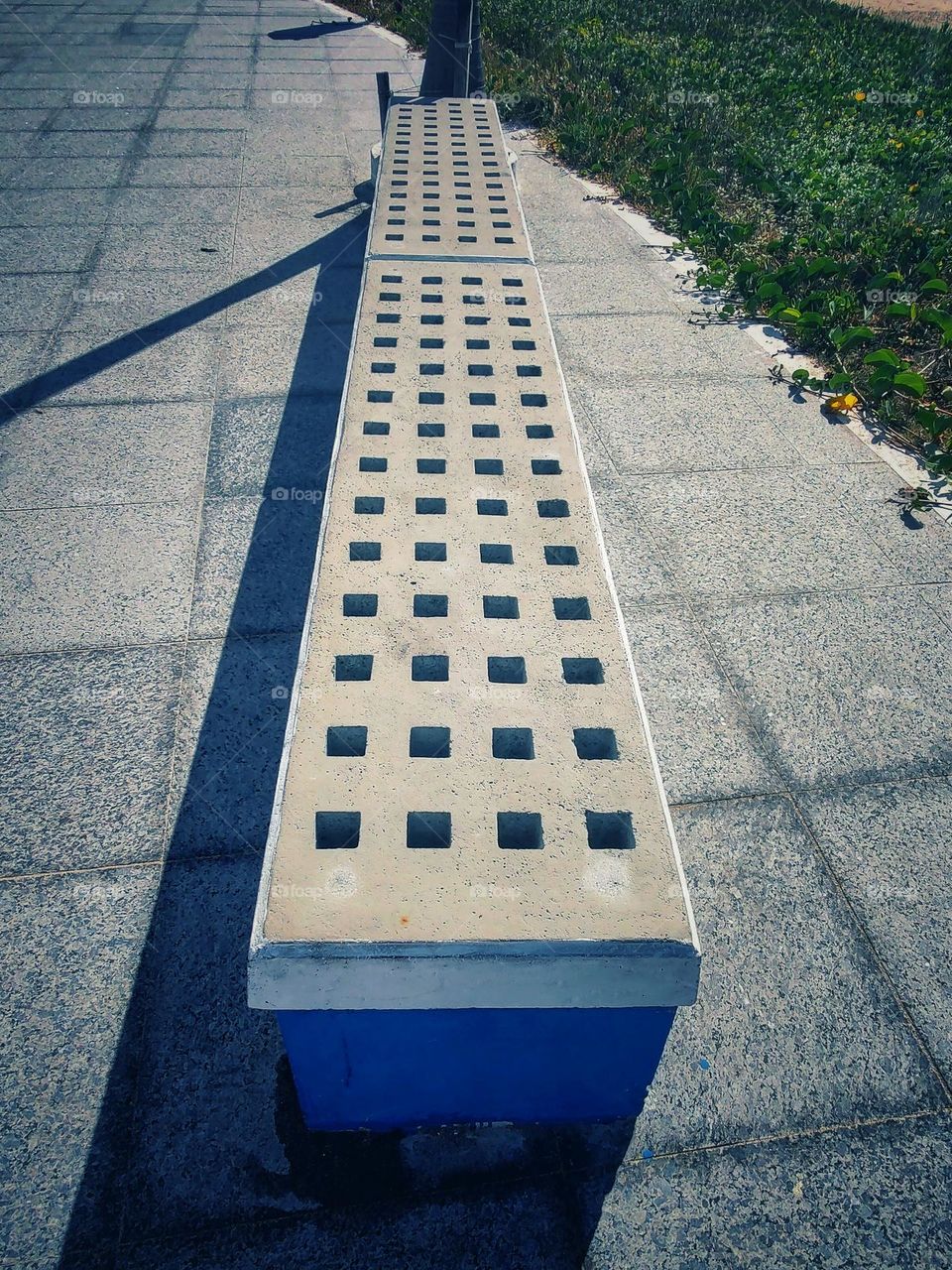 a rectangular bench on the beach promenade made of cement, with the seat filled with hollow squares to avoid any accumulation of water when it rains.