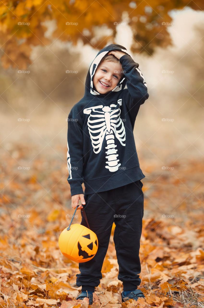 Cute and funny little boy in spooky skeleton costume for Halloween carnival, hold pumpkin bucket for sweets 