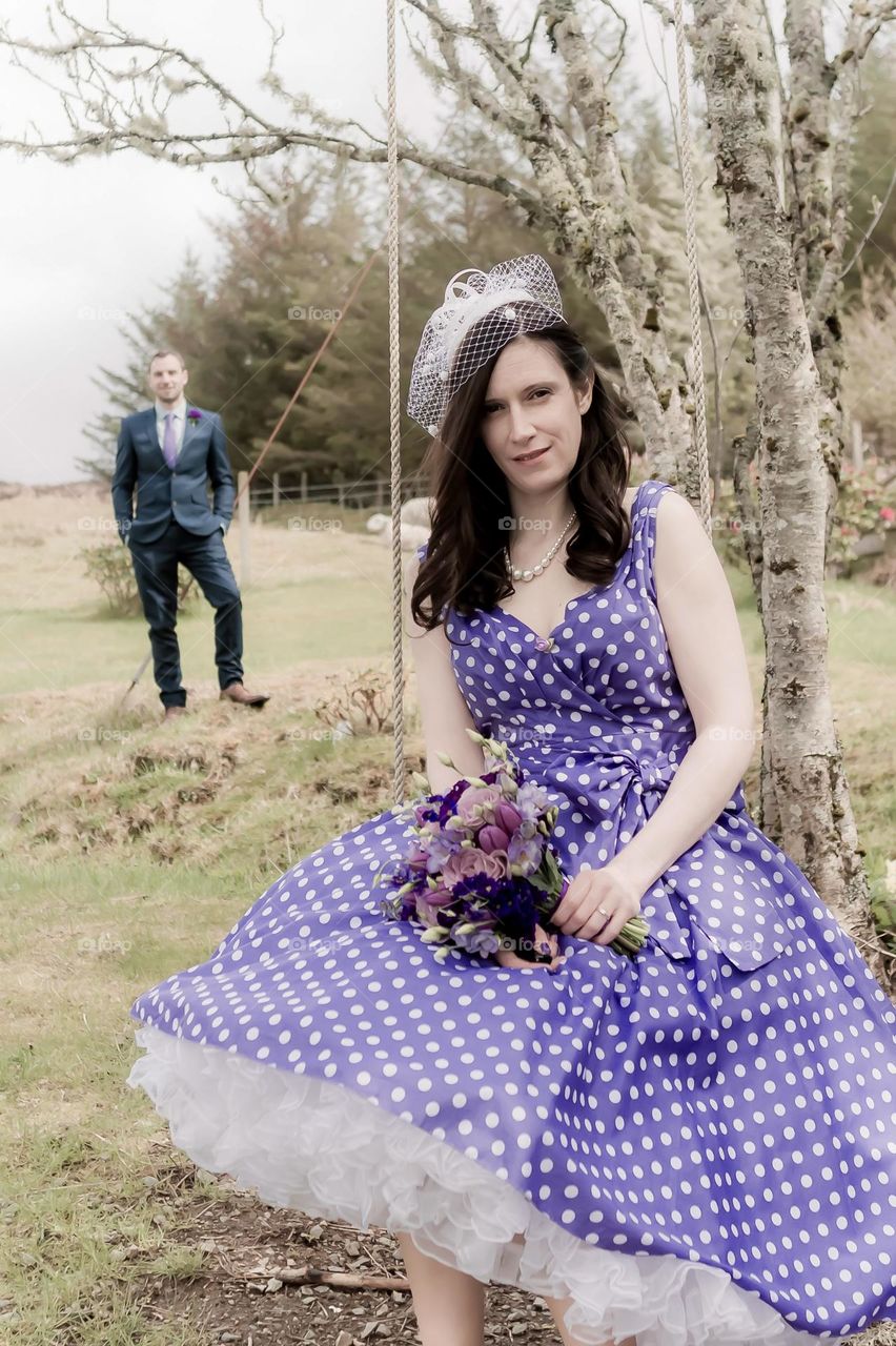 A woman in a purple polka dot dress sits on a swing as a man in a teal suit waits in the distance