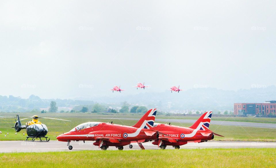 Two Red Arrow Hawk T1A standing on runway, whilst three Red Arrows take off in background.