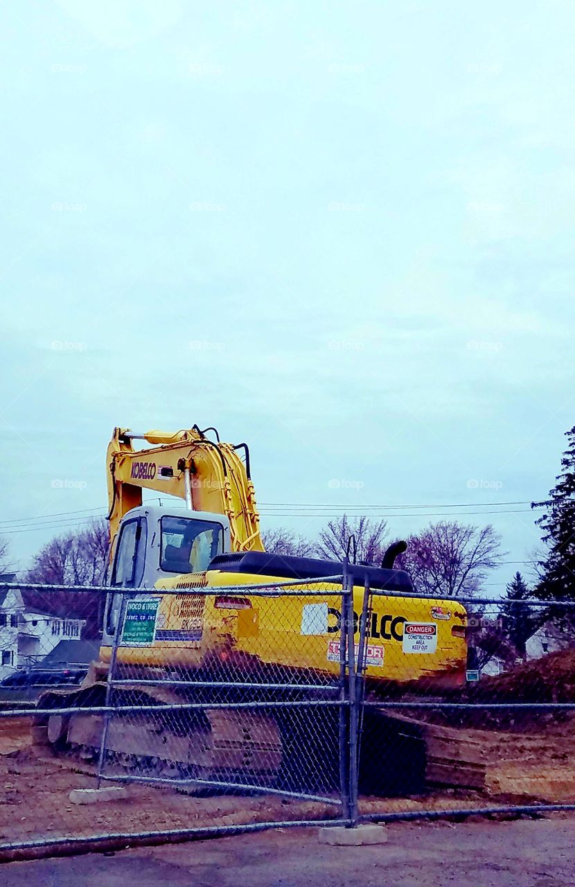 Big tractor with scoop moving earth & rocks, trees look tiny next to it. Its scoop was huge to clear the land away on this warm sunny day.
