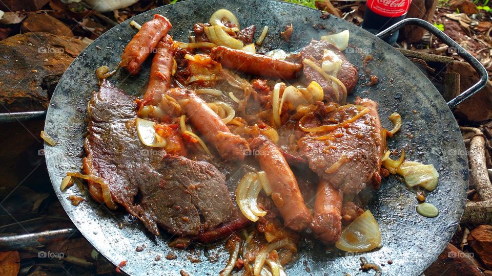 Carnes bovinas e suínas assadas no disco de grade, arado de trator, com fogo de álcool. Alcatra e linguiça com sal, tomate, cebola, pimenta, limão e queijo.