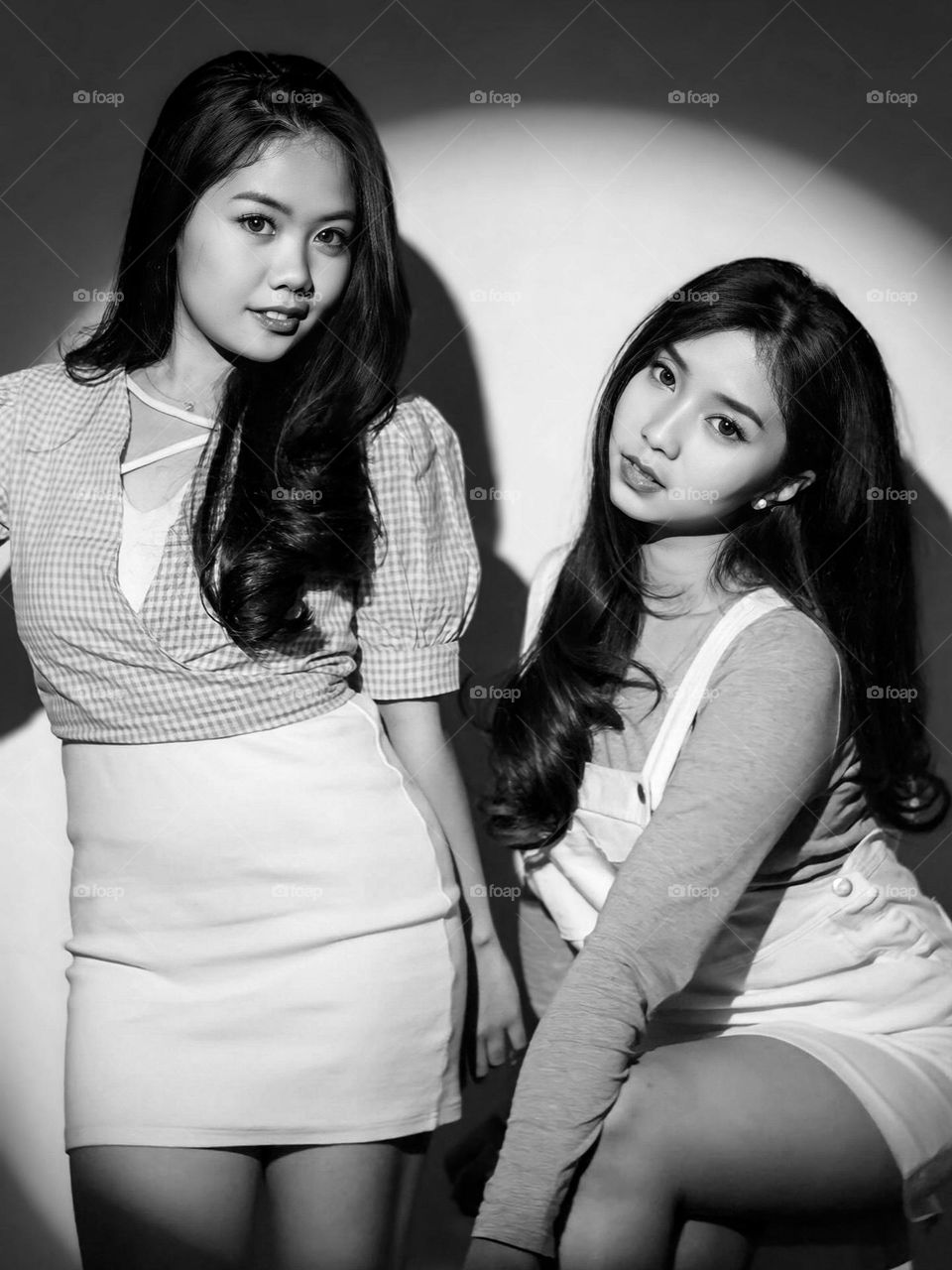 Portrait of two young women with their beautiful faces posing in front of a neutral background. They wore black and white clothes, one wore a top and skirt, while the other wore a dress.