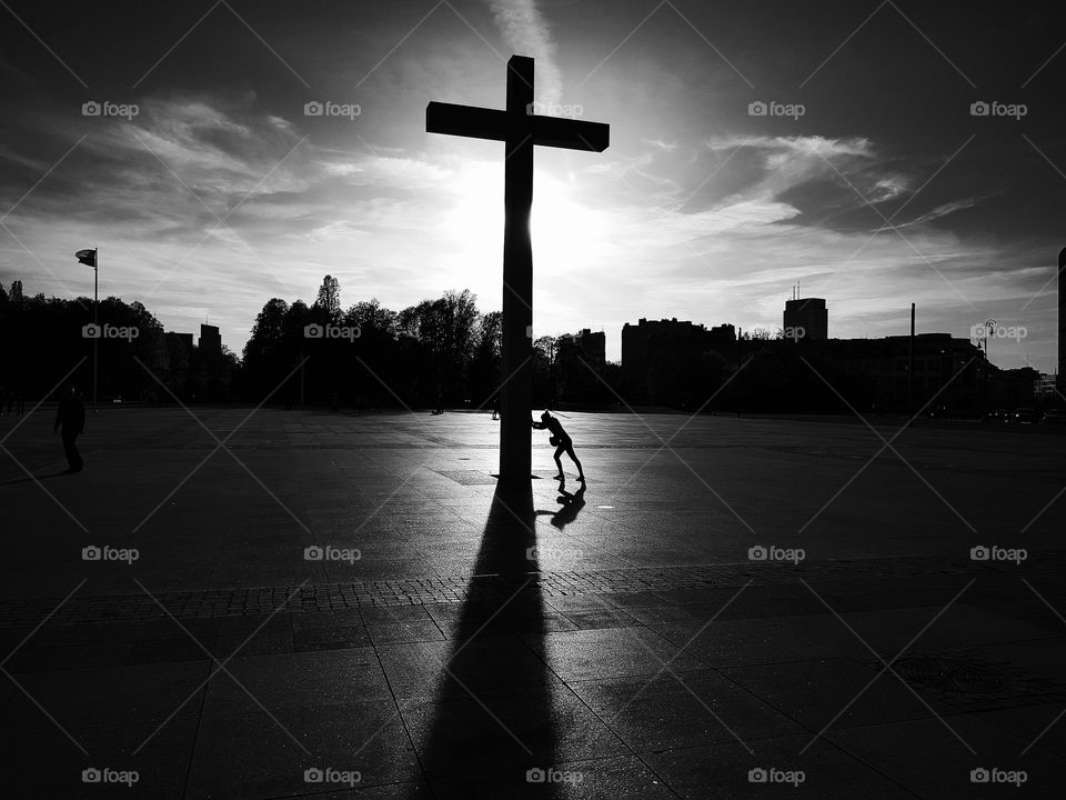 Silhouette of a cross monument and a person stretching on it making look like they are pushing the giant object. Warsaw. Monochrome.