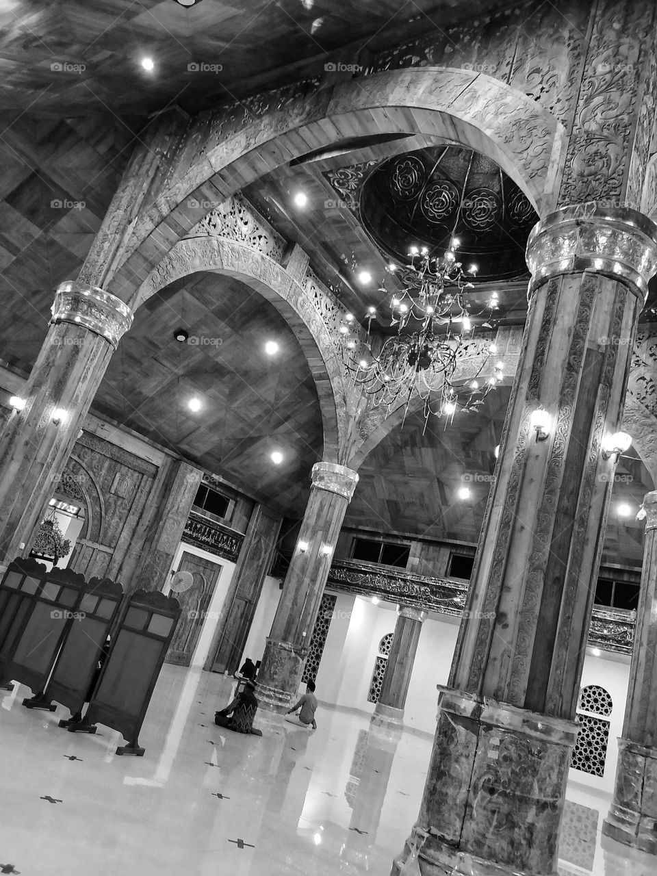 The interior of a magnificent mosque in black and white effect, carved wooden pillars, luxurious chandeliers, white tiled floors and several people praying.