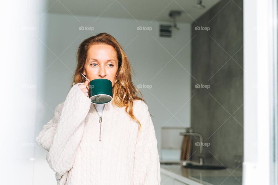 Young adult forty years beautiful blonde woman with long hair using smartphone and drinking morning tea on the kitchen at home