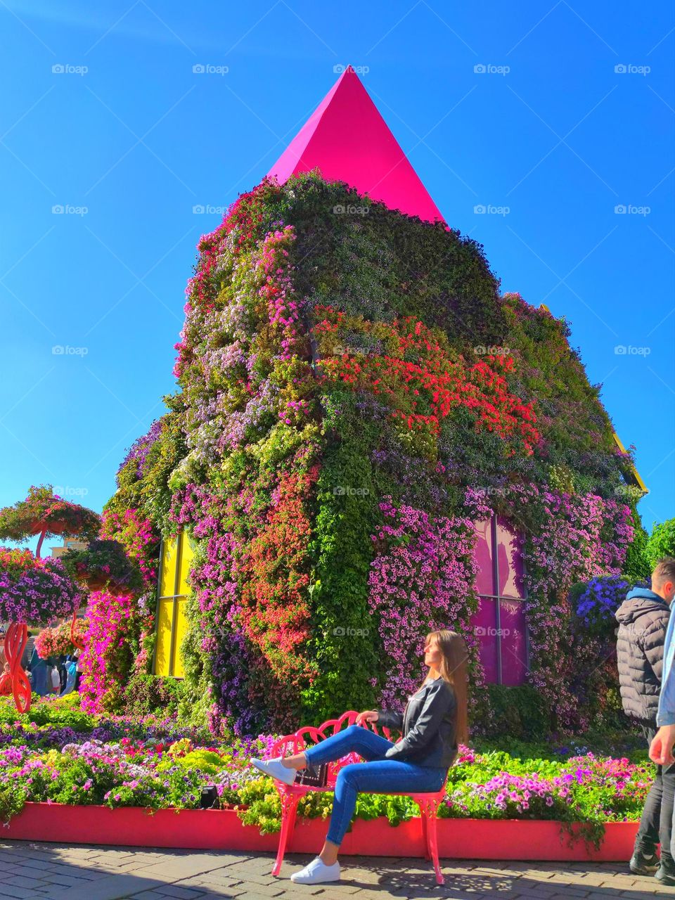 Plants.  The house is completely overgrown with colorful flowers.  Blue sky on the background of the house.  A girl sits in the foreground and basks in the sun