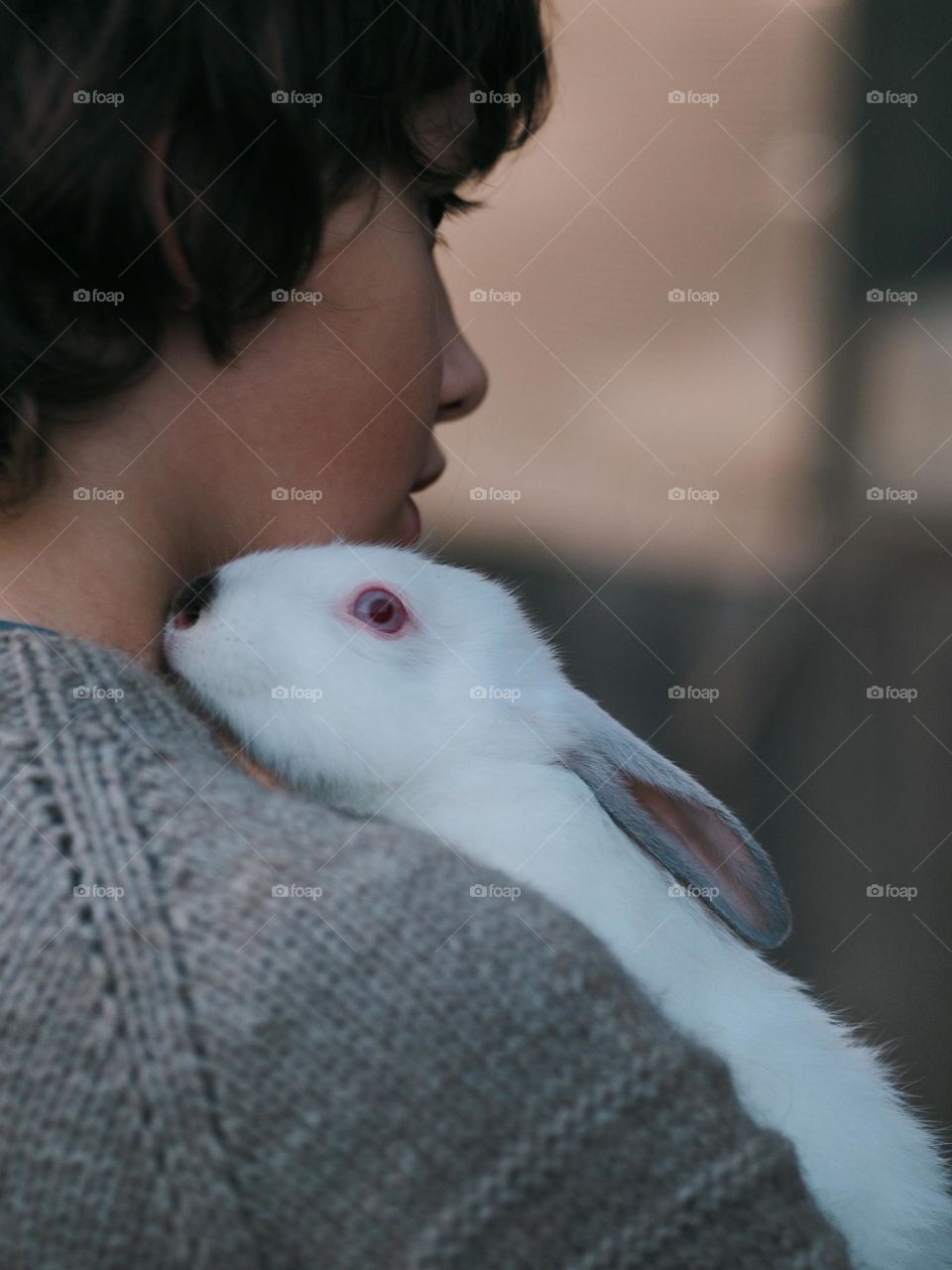 A cute little boy with dark hair in a gray knit sweater is holding a little white rabbit, happy Easter, friendship and love concept 