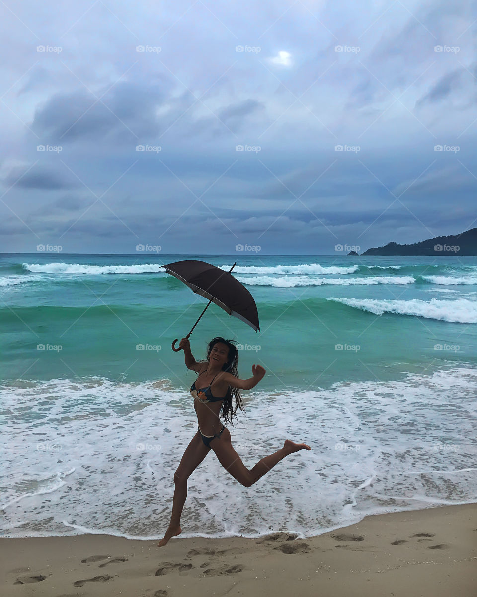 Young woman happily jumping above the ocean waves with umbrella during the stormy weather 
