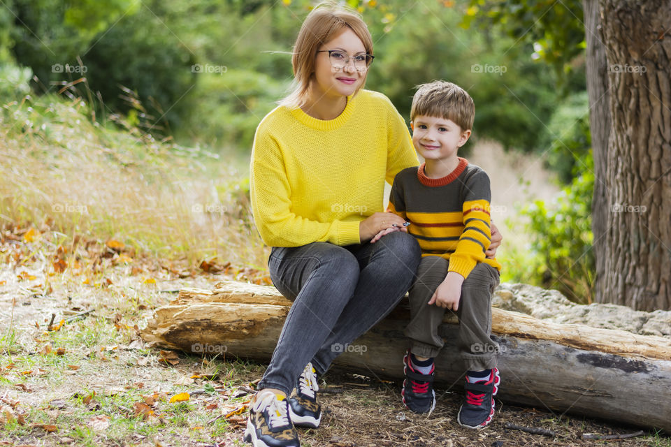 Mother and son in a cozy yellow brown sweater are sitting in the forest in autumn on a sunny day smiling looking straight into the camera picnic rest on nature weekend